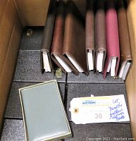 Box containing 7 Bugatti leather agenda pocket books in multiple brown shades and 1 gray mini address book on top, along with multiple black boxes beneath.