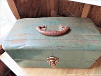 Closed view of vintage green metal toolbox with brown handle and latch showing some wear and rust.