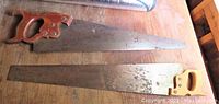 Two vintage hand saws with wood handles laid side by side on a wooden surface; top saw has darker brown handle, bottom one lighter handle.