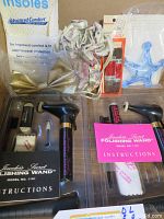 Two vintage Jeweler's Secret Polishing Wand kits in original plastic containers with polishing cloths, brushes, and instruction manuals. Additional miscellaneous items include shoe insoles, a gold bow, floral drawstring bag, and blue plastic tool.