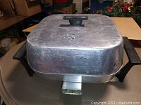 Overview photo showing the aluminum rectangular vintage electric skillet with black handles and vented lid, placed on a surface.
