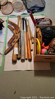 Photo showing six baseball bats, two wooden boomerangs, baseball gloves, catcher's mask and colorful baseballs in a box.