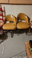 Two vintage swivel chairs with mustard yellow vinyl upholstery, curved backrests with open space below, and metal bases with four legs visible.