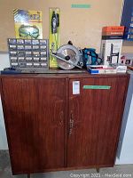 Wooden cabinet with doors and hardware on top including Makita circular saw, Black & Decker Guidemate, safety goggles package, and hardware organizer box