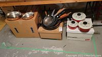 Photo showing a grouping of red pots with white lids and a stack of various frying pans placed on cardboard boxes in a basement setting.
