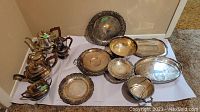 Overview of the silver plated tea set and serving dishes laid out on white cloth showing teapots, trays, and bowls