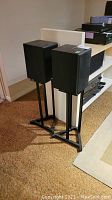 Pair of black Paradigm bookshelf speakers on black metal stands, approximately 8x12x37 inches in size, placed on a carpeted floor indoors.