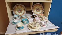 Overview of two decorative plates behind ten assorted tea cup and saucer sets