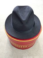 Top angled view of black fedora hat resting on original red and yellow Christys London branded box.