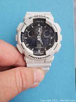 Front view of white Casio G-Shock watch showing analog hands, digital displays, and button labels: Adjust, Mode, Reverse, Forward, Protection, Shock Resist.