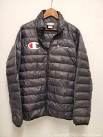 Front view of black men's puffer jacket with Champion logos on chest, hung on wooden hanger against white wall.