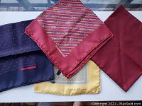 Four men's silk pocket squares laid flat: navy with red anchors (Hermès), cream with blue/yellow motif (Salvatore Ferragamo), striped red tones, and solid deep red.