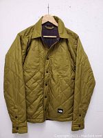 Men's North Face men's reversible quilted jacket hanging on wooden hanger against white wall. Shows olive green diamond stitched side with two chest pockets and button closure.