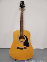 Full front view of the acoustic guitar showing natural wood finish, black pickguard, six tuning pegs, dot inlays on fretboard, and missing three strings.