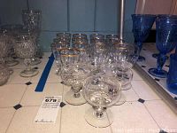 Clear cordial glasses with gold rims arranged in rows with smaller dessert bowls and glasses to the front and right, all glassware is clear with gold detailing on cordial glasses.