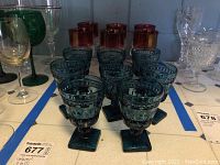 Front view of goblets including six blue pressed glass goblets and cranberry two-toned goblets behind them, along with green and clear glassware.