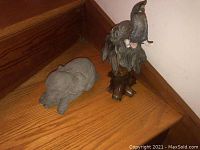 Two elephant statues side by side, one gray cement and one painted plastic on wooden base.