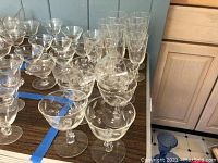 Wide view of multiple etched glassware items on wood table showing seven dessert bowls, various glasses with etched floral patterns.