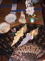 Overview of lot showing figurines including baby carriage with baby, glass frog, angels, small doll figure, snowman pin, and fabric hand fan beneath