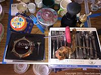 View of boxed First Impressions plastic flatware sets, vintage pepper grinder, slap-chop, ice cream pot, various mugs, Bombay wine stopper and coasters, Simpsons coasters.