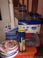 Lot overview showing stacked paper cups, paper plates, Michael Graves blue food storage containers, various sized containers with blue lids, and packaging box for Reynolds Cleverware containers.