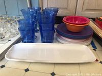 Photo showing blue plastic cups, clear goblets with gold rims, plastic bowls in orange and pink, stack of plastic plates, and a long white plastic tray.
