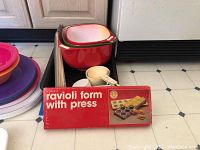 Photo showing red Rowoco ravioli form with press in original packaging alongside various plastic nesting bowls and kitchen tools.