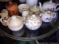 Overview of the lot showing wooden mug, floral and white ceramic teapots, sugar bowls, and creamer on glass table.