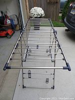 Top-down view of metal drying rack fully extended on driveway, showing multiple horizontal rods and foldable legs.