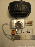 Silver Nikon Coolpix LS digital camera with lens open, wrist strap, Panasonic AA rechargeable batteries, Lowepro case, 2GB SD card, stainless steel ruler for scale, white background.