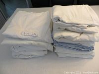 Nine white and light-colored flat queen sheets in two piles on a dark surface, showing clean and folded condition.