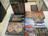 Eight assorted puzzles on a table including 3D Cologne Cathedral box, panoramic 12th Day of Christmas, Ravensburger village scene, Eurographics antique world map, and several others.