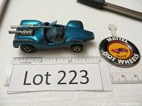 Photo of teal metallic Hot Wheels car with open windows and badge for scale and identification.