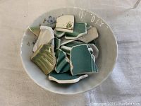 Close-up image of a white bowl with ceramic sea glass shards in various shades of green, teal, and white, showing the textures and glazes of the shards.