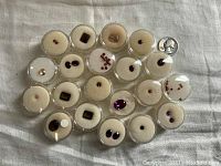 Top-down view of 25 individual faceted gemstones placed in circular plastic containers on cream felt pads showing a variety of sizes and colors with a quarter for size reference.