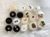 Top view of various faceted and cabochon stones in small plastic containers arranged in rows on a beige surface, showing a wide range of colors and cuts including heart-shaped, oval, round, and rough geode.
