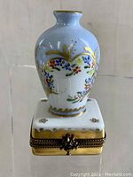 Hand painted porcelain pill box with light blue miniature vase lid featuring floral garlands and gold detailing, showing front view.