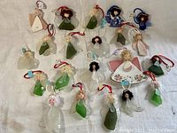 Overall grouping of 21 sea glass angel ornaments laid out on white fabric background showing variety of greens, clear glass, and some decorated dresses with hanging loops