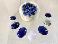 Top-down photo showing multiple polished lapis lazuli cabochons and one turquoise cabochon, arranged on a reflective surface with a white padded container for smaller stones.