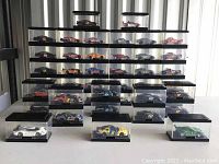 Wide front view showing 28 diecast cars in individual clear display cases arranged in stacked rows and on table