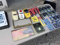Full overview of the lot displayed on a table: three car models, Porsche metal frame, Shield license plate holder, Plate Guard package, and multiple auto emblems sealed in packaging.