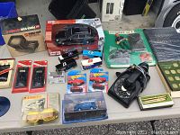 Overview of model cars including Maisto black car, Hot Wheels cars, packaged police van, model car parts, unopened darts, boxed binoculars or goggles, vintage rotary dial phone, and Cross pens boxed set.