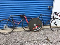 Two black carbon bike frames side by side against blue door, one with red fork and one mixte-style top tube, and three wheels (two spoked, one disc) mounted