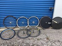 Eight bicycle wheelsets including disc wheels, freewheel-equipped rims, and clincher wheels displayed against blue roll-up door