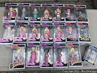Overview of 24 Rock Candy dolls in unopened boxes displayed on surface showing Barbie, Harley Quinn, Batgirl, and Supergirl dolls in various outfits.