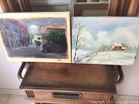 Photo showing two paintings on canvas resting on a metal cabinet, one depicting a street scene with buildings and people, the other a winter landscape with snow and a small house.