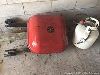Red rectangular BBQ grill cart with wheels and small white propane tank beside it close to wall.