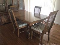 Photo showing the wooden dining table along with four chairs, placed on hardwood flooring with a china cabinet in the background.