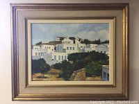 Framed painting showing white building structures and surrounding greenery in a landscape format with beige/gold frame.