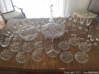 Wide view of all crystal and glassware items arranged on a wooden table including bowels, glasses, decanter, and pedestal dish.